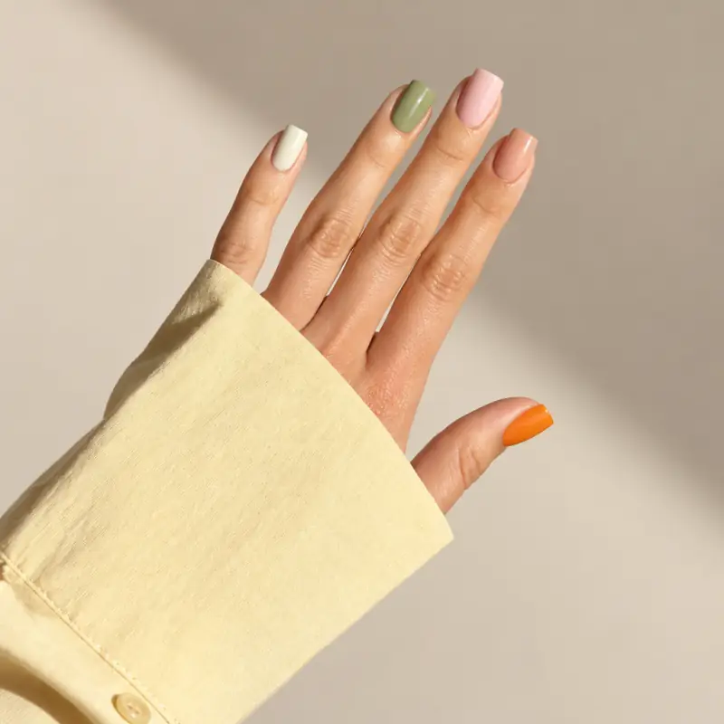 A hand in a butter-yellow sleeve with square nails in cream, sage, blush, nude and tangerine, in afternoon sunlight